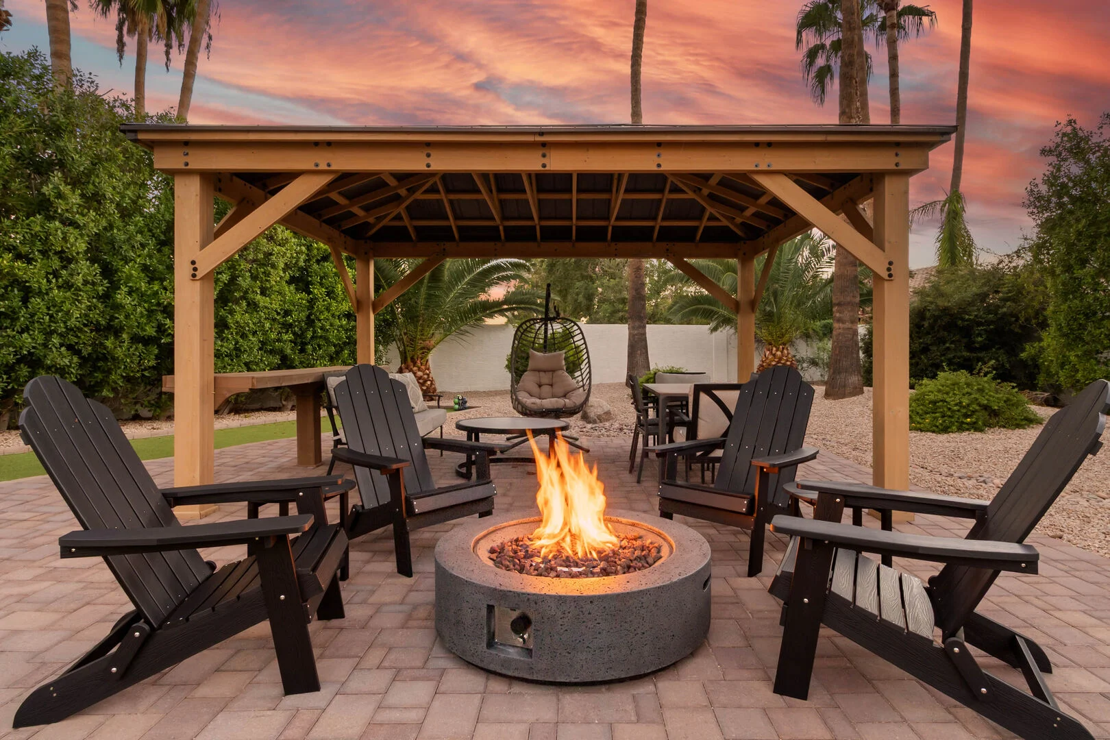 Cedar gazebo over a paver patio with a round gas fire pit and lounge chairs at sunset